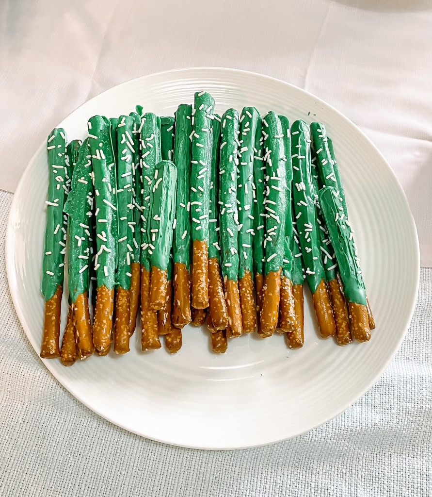 A white plate with pretzel rods dipped in green chocolate with white jimmy sprinkles that resemble cacti
