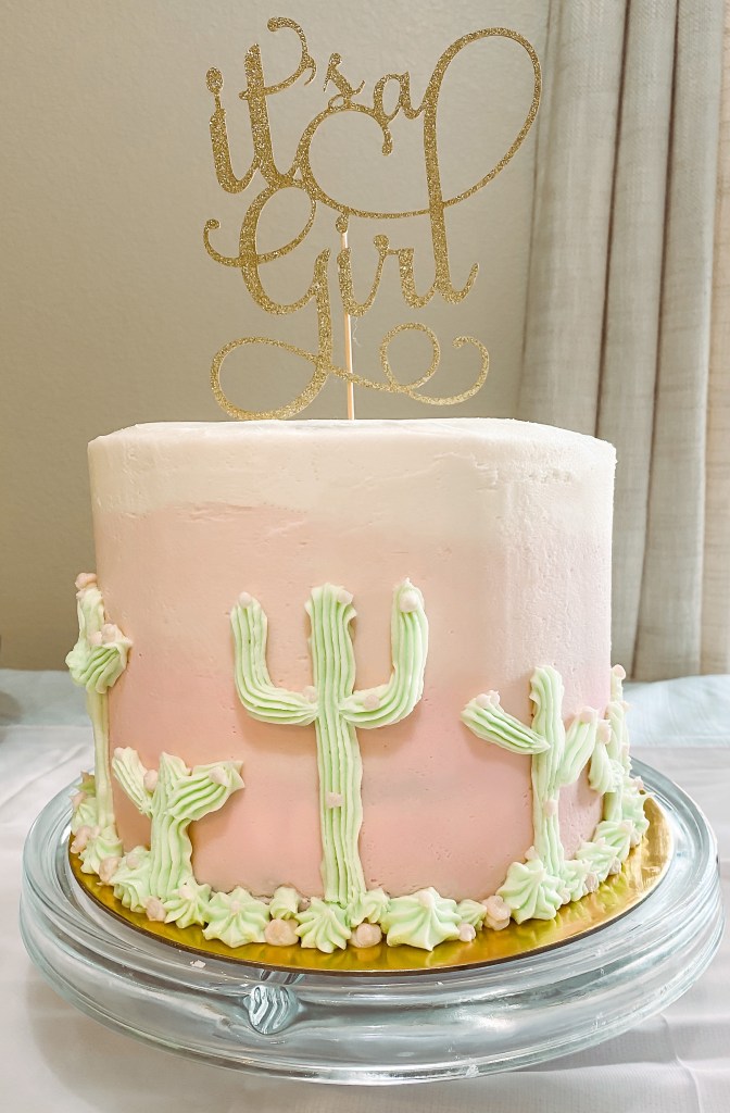  A pink and white ombre cake with green cacti and succulents, with a sparkly gold "It's a Girl" cake topper.
