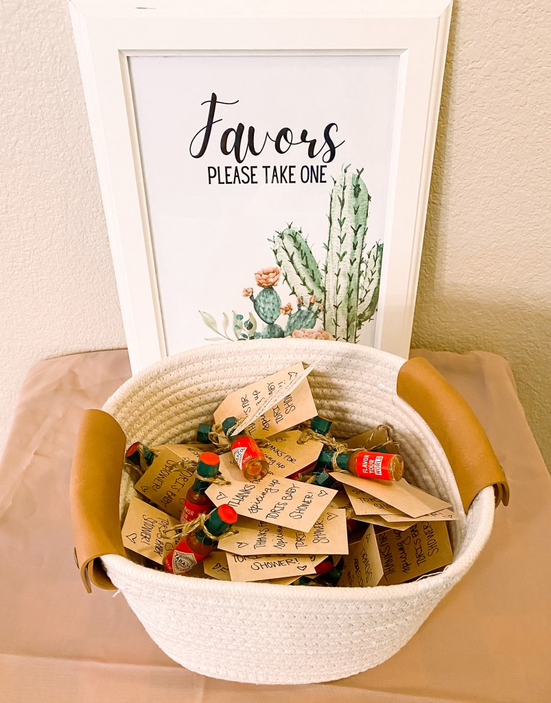 A basket full of mini bottles of hot sauce for baby shower favors with a tag that reads "Thanks for spicing up the baby shower." The basket is on a small table with a sign by it that reads "Favors, please take one."