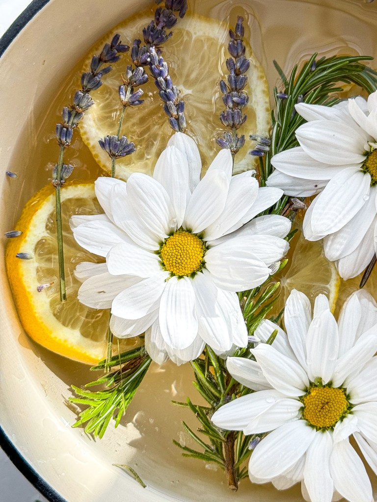 A close up view of a enamel cast iron pot filled with water, lemon slices, lavender, rosemary, and white daisies to create a fragrant simmer pot.