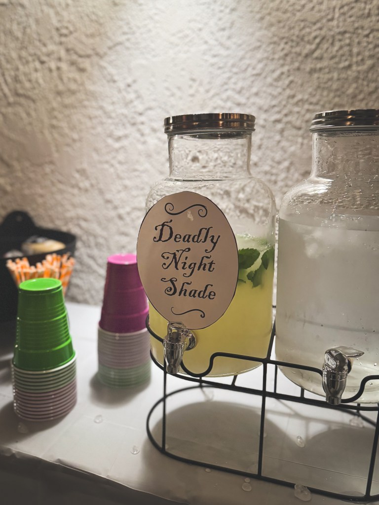 A drink dispenser holding a light green liquid with mint leaves with a sign that reads "Deadly Night Shade."