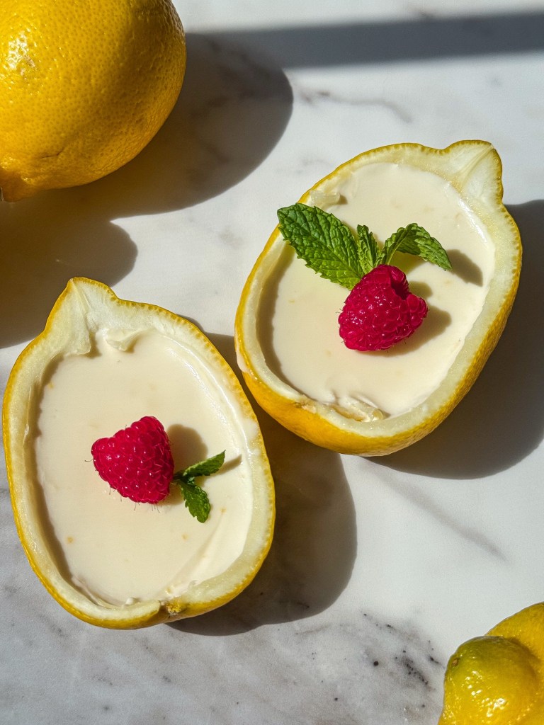 Creamy Lemon Posset, a classic British dessert, being served in hollowed-out lemon halves, with a vibrant raspberry and sprig of mint as garnish.
